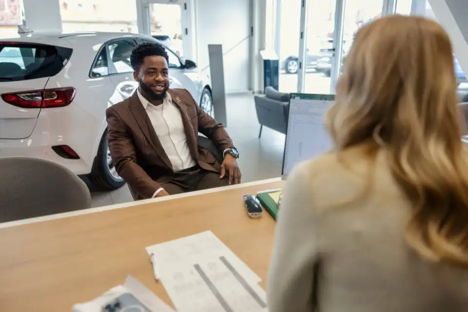 Man in a brown suit smiles while seated at a desk across from a female staff member in a car showroom with a white car in the background.